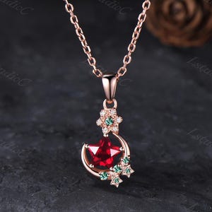 May include: A rose gold necklace featuring a pendant with a central red star-shaped gemstone nestled within a crescent moon frame. The design is accented with small clear and green stones arranged in star shapes along the crescent and at the top bail.