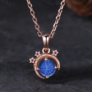 Peut inclure: Un collier en or rose avec un pendentif orné d'une pierre bleue ronde à facettes. La pierre est entourée d'un croissant de lune décoré de petites étoiles, dont deux sont serties de minuscules pierres rouges. Le pendentif est sur une chaîne.