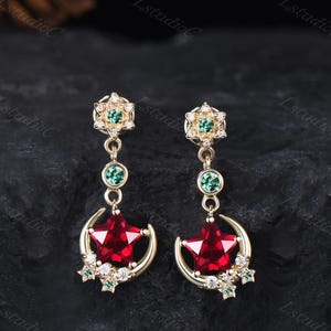 Puede incluir: Un par de pendientes colgantes dorados con un poste en forma de estrella con una piedra verde, un conector circular verde y un dije con una gran estrella roja dentro de una media luna, decorado con pequeñas piedras transparentes.