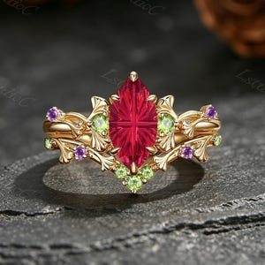 May include: A gold ring featuring a central marquise-cut red gemstone with a faceted surface. The band is designed with intricate leaf-like metalwork, accented by small round green and purple gemstones embedded along the decorative branches.