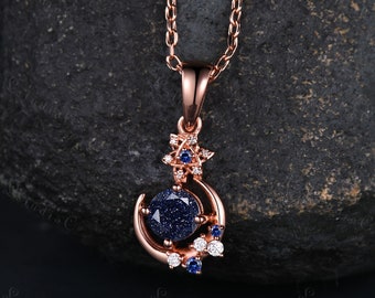 May include: A rose gold pendant necklace featuring a large, round, dark blue glittery sandstone center stone set within a crescent moon frame. The design includes a small star accent at the top and clusters of tiny clear and blue stones along the side.