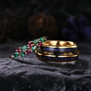 May include: Two rings, one with a gold band and a dark inlay with blue and gold flecks, the other with a thin band and a design of green and red gemstones. The rings are displayed on a dark, textured surface.