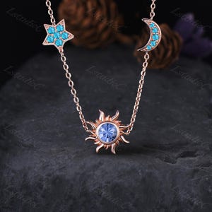 May include: A rose gold necklace with celestial-themed pendants. The necklace features a star, a crescent moon, and a sun, each embellished with turquoise gemstones. The sun pendant has a central blue gemstone. The necklace is displayed on a dark, textured surface.