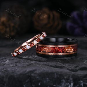 May include: A rose gold band featuring alternating white opal and red garnet stones, paired with a wider black band featuring a central inlay of crushed red stone and copper-colored flakes bordered by brushed rose gold metal edges.