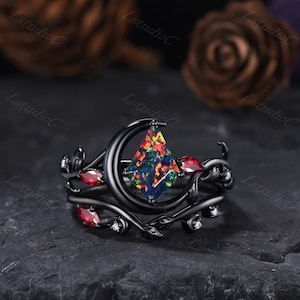 May include: Black ring set featuring a central, multi-colored gemstone in a diamond shape, framed by a black crescent moon. The ring is adorned with red gemstones and delicate leaf and vine details, with small white accents.