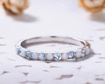 May include: A silver-toned band ring featuring an alternating pattern of round, iridescent white opals and clear, faceted blue gemstones set in a row along the top half of the metal band, creating a delicate and symmetrical jewellery piece.