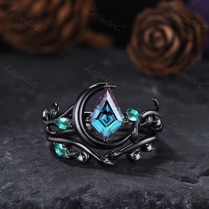 Op de afbeelding: Zwarte ringenset met een centrale, vlieger-vormige, teal en paarse edelsteen. De ring is versierd met groene edelstenen en kleine witte diamanten, gezet in een zwart, bloemig ontwerp. De ring heeft een halve maan vorm.