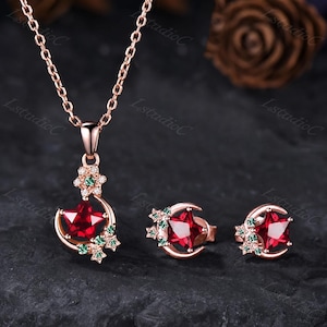 May include: A rose gold necklace and matching stud earrings set. Each piece features a central red star-shaped gemstone, accented by a crescent moon frame and smaller green and clear stones arranged in a cluster around the star.