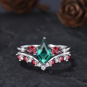 May include: A silver ring set with a large emerald-cut green gemstone and smaller red and white gemstones. The ring set has a crown-like design. The gemstones are set in a decorative pattern.