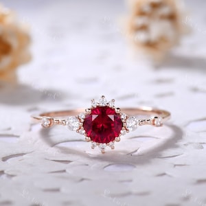 May include: A rose gold engagement ring with a large, round, red ruby gemstone surrounded by a halo of smaller, white diamonds. The band is delicate and features additional small diamonds.
