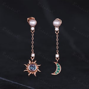 Puede incluir: Un par de pendientes colgantes con una perla en la parte superior y un dije de sol y luna en la parte inferior. El dije de sol tiene una piedra preciosa azul en el centro y el dije de luna tiene piedras preciosas verdes.