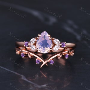 May include: A rose gold engagement ring set with a pear-shaped pale pink gemstone and smaller clear gemstones. The ring is accented with delicate leaf designs and small purple gemstones.