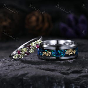 May include: Two rings, one with a silver band and a row of green and red gemstones, the other with a black band, blue and green inlay, and gold accents. The rings are displayed on a dark, textured surface.