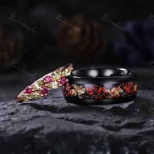 Puede incluir: Dos anillos: una banda de oro rosa con piedras preciosas verdes y rosas, y un anillo negro con motas rojas y doradas. Los anillos se colocan sobre un fondo oscuro y texturizado.