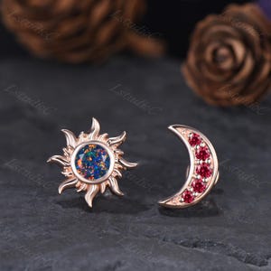 May include: A pair of celestial-themed earrings. One earring is a sunburst design with a central opal stone. The other earring is a crescent moon shape, adorned with red gemstones. Both earrings have a rose gold finish.