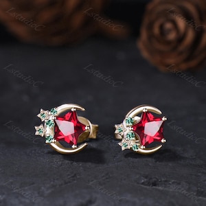 May include: A pair of gold-toned stud earrings featuring a crescent moon shape accented with a central star-cut red gemstone and smaller green gemstone accents arranged in a cluster along the side of the moon and star design.