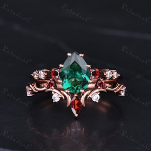 Könnte beinhalten: Ein Verlobungsring mit einem birnenförmigen smaragdgrünen Edelstein, der mit einem passenden Band kombiniert wird. Das Band hat ein zartes Blumenmuster mit roten Edelsteinen und weißen Diamanten.