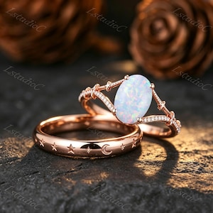 Könnte beinhalten: Ein Ring-Set aus Roségold mit einem oval geschliffenen Opal als Mittelstein auf einer Ringschiene mit kleinen Sternen und klaren Steinen, dazu ein passender Ring aus Roségold mit eingraviertem Muster aus Sternen und einem Halbmond.