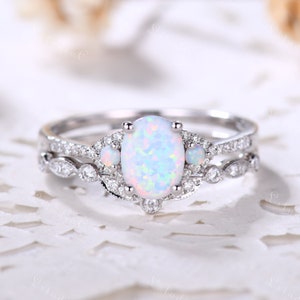 May include: A silver-toned ring featuring a large oval-cut iridescent opal center stone flanked by two smaller round opals. The band and setting are embellished with numerous tiny clear pavé stones arranged in a delicate, intricate pattern.