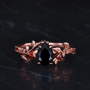 May include: A rose gold engagement ring with a pear-shaped black diamond and red gemstones set in a delicate vine design.