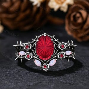 Puede incluir: Un anillo de metal negro con una gran piedra central roja de corte ovalado, rodeada de piedras rojas más pequeñas en engastes con forma de rosa. La banda presenta protuberancias en forma de espinas y ópalos blancos ovalados y lisos.