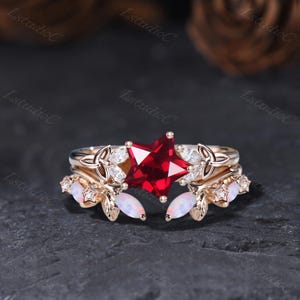May include: A rose gold ring featuring a central star-shaped red gemstone flanked by marquise-cut clear stones. The band includes leaf-like metalwork, small star accents, and oval-shaped iridescent stones arranged in a botanical-inspired design.
