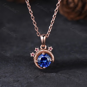 May include: A rose gold necklace featuring a circular pendant with a central round-cut deep blue gemstone. The pendant is framed by a crescent moon shape adorned with small star motifs, each accented with a tiny, vibrant red gemstone inlay.