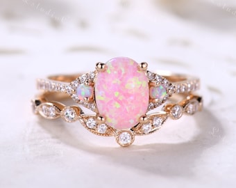 May include: A rose gold ring set featuring a large oval pink opal center stone flanked by two smaller round opals. The band and the curved nesting band are accented with numerous small clear stones set in a delicate, intricate metal design.