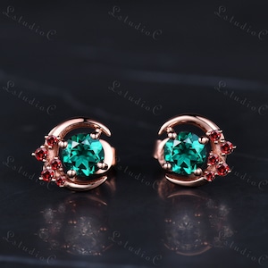 May include: A pair of rose gold earrings with a crescent moon design. Each earring features a round green gemstone surrounded by smaller red gemstones.