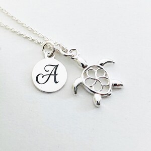 Puede incluir: Collar de plata con un pequeño dije de tortuga y un dije redondo con la letra A grabada.