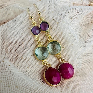 Könnte beinhalten: Goldene Ohrringe mit drei Edelstein-Tropfen. Der obere Tropfen ist lila, der mittlere Tropfen ist grün und der untere Tropfen ist rot.