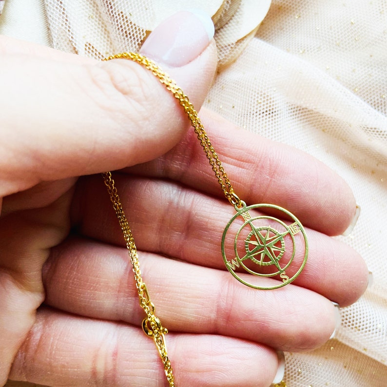 Pu&ograve; includere: Una collana con catena dorata e un ciondolo a forma di bussola. La bussola &egrave; un cerchio con una stella al centro e le lettere N, S, E e W attorno al bordo.