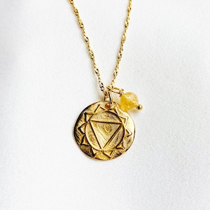 Puede incluir: Collar de oro con un colgante redondo que presenta un símbolo de chakra detallado y un pequeño amuleto de piedra amarilla.