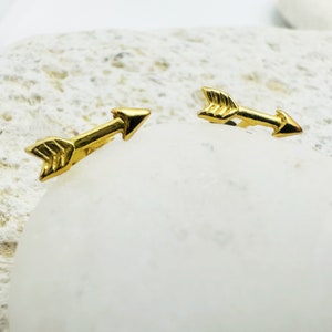 Puede incluir: Un par de pendientes de aro de flecha de oro. Las flechas son simples y minimalistas en diseño.