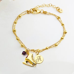 Op de afbeelding: Gouden kettingarmband met een bedel in de vorm van een gouden schaats, een klein paars kristal en een gouden schijf met de letter 'A' erop gegraveerd.