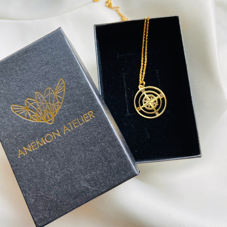 Pu&ograve; includere: Una collana con ciondolo a forma di bussola dorata in una scatola nera con il testo "ANEMON ATELIER" sulla scatola.