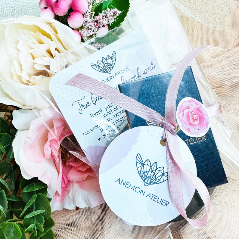 Puede incluir: Una peque&ntilde;a caja de regalo envuelta en una cinta rosa con una etiqueta blanca que dice "True beauty" y "Thank you for being part of... This is a promise... No words can express... was more... beyond words". La caja est&aacute; rodeada de flores rosas y una bolsa de pl&aacute;stico transparente.
