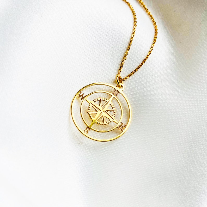 Pu&ograve; includere: Una collana d'oro con un ciondolo a forma di bussola. La bussola presenta un design dettagliato con le lettere N, S, E e W.