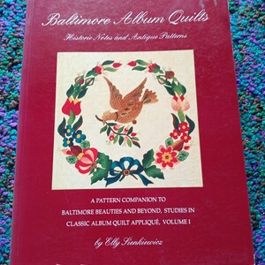 May include: A red book titled "Baltimore Album Quilts" with the subtitle "Historic Notes and Antique Patterns." The cover features a central square image of a bird holding a sprig, surrounded by a floral wreath. The author's name is Elly Sienkiewicz.