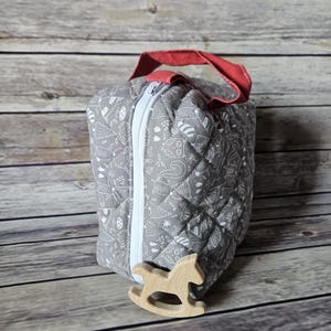 Puede incluir: Una bolsa acolchada gris con cremallera blanca y asa roja. La bolsa presenta un patrón repetido de ilustraciones blancas con temática de bosque. Un pequeño juguete de madera con forma de caballito balancín está delante de la bolsa.