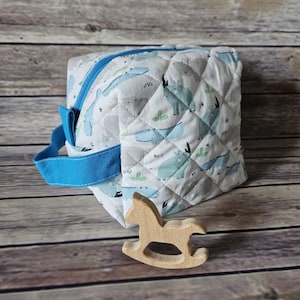 Puede incluir: Un bolso de tela acolchada blanca con cremallera y asa azules, estampado con motivos de dinosaurios azul claro. Un pequeño juguete de caballito balancín de madera está junto al bolso. El bolso mide aproximadamente 15 cm de ancho y 13 cm de alto.