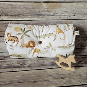May include: A white baby changing mat featuring a jungle animal design with tigers, lions, and crocodiles. A small wooden rocking horse toy is next to the mat. The mat is folded and secured with a button.