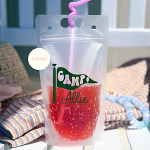 May include: A clear, flexible plastic drink pouch filled with a red liquid and a pink straw. The pouch features a green pennant flag graphic with the text "CAMP" and the name "Allie" in green script. A circular white logo reads "POSIE MAE".