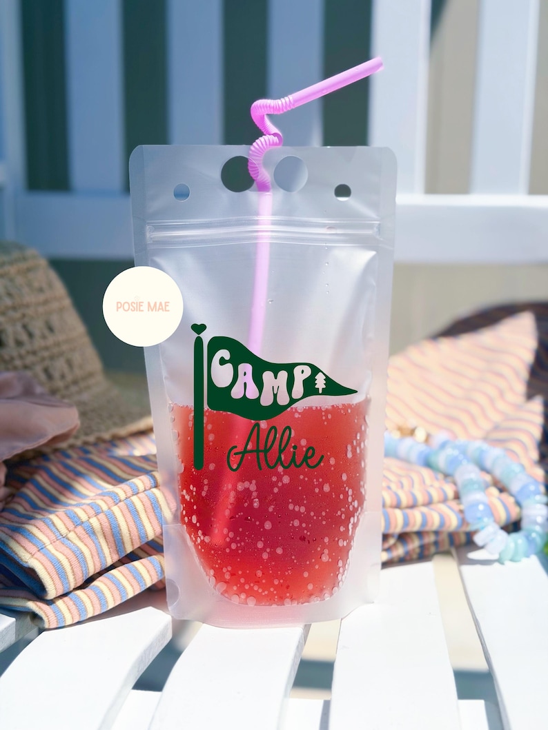 May include: A clear, flexible plastic drink pouch filled with a red liquid and a pink straw. The pouch features a green pennant flag graphic with the text "CAMP" and the name "Allie" in green script. A circular white logo reads "POSIE MAE".