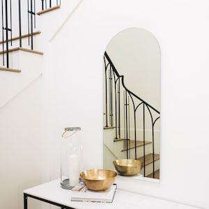 May include: A modern entryway scene featuring a white marble-topped console table with a black metal frame. The table holds a gold hammered bowl, a glass lantern with a candle, and a stack of books. An arched mirror reflects a staircase.
