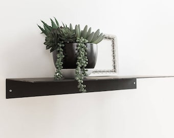 May include: A black metal shelf with a succulent plant in a black pot. The plant has green leaves and hangs down from the pot. The shelf is mounted on a white wall.