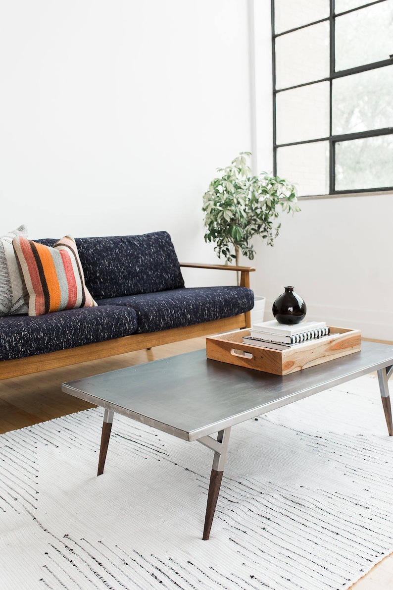 May include: A dark blue sofa with wooden arms and a striped pillow sits in front of a large window. A metal coffee table with wooden legs sits on a white and black patterned rug. A wooden tray with books and a black vase sits on the coffee table.