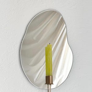 Straight shot of the wall mounted organic shape Mar Mirror Candle Sconce or Mirror Candle Holder. The candleholder is gold & holding a green unlit candle. The reflection is of the gold candle holder, the green unlit candle & the cream background.