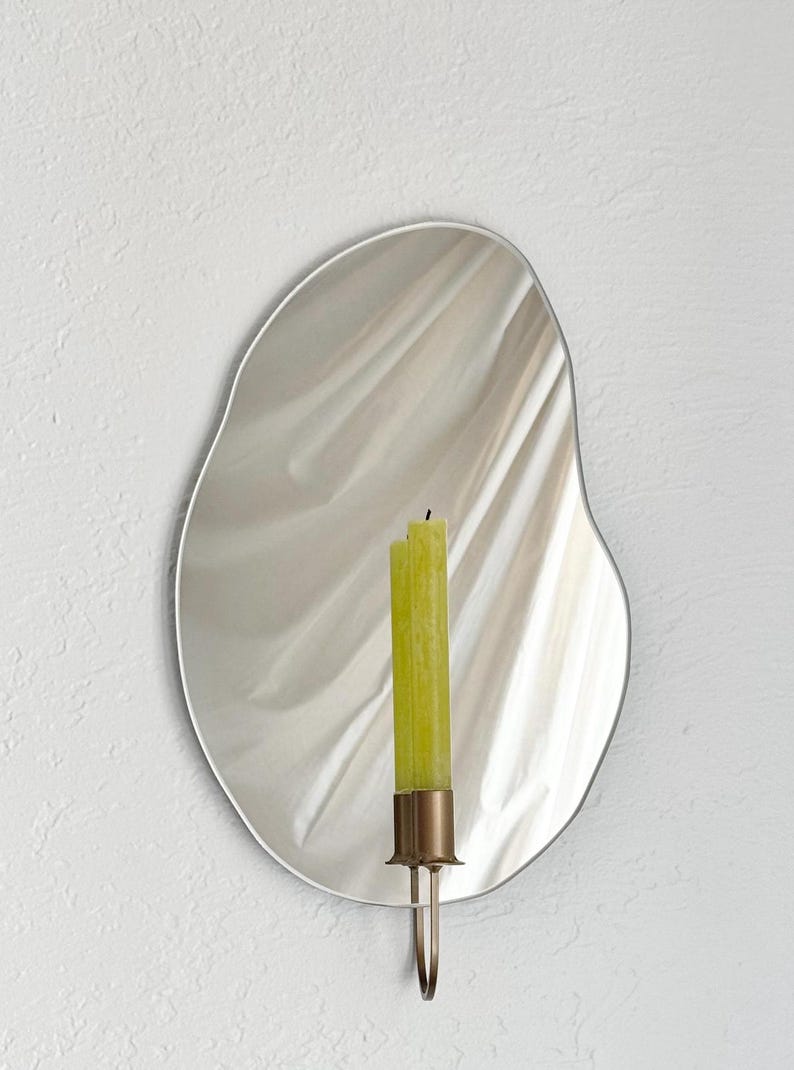 Straight shot of the wall mounted organic shape Mar Mirror Candle Sconce or Mirror Candle Holder. The candleholder is gold & holding a green unlit candle. The reflection is of the gold candle holder, the green unlit candle & the cream background.