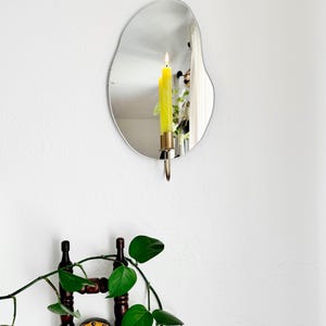 the wall mounted organic shape Mar Mirror Candle Sconce or Mirror Candle Holder. The candleholder is gold & holding a green lit candle. The reflection is of the gold candle holder, the candle & the cozy home setting with plants and decor.
